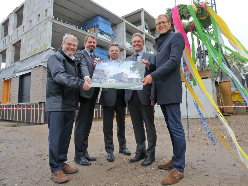 F&uuml;nf M&auml;nner halten ein Bild in die Kamera vor einem Rohbau, dahinter ein Richtkranz.